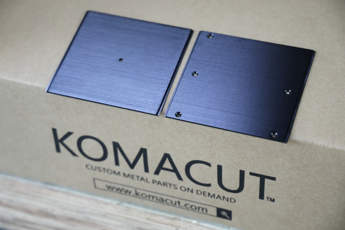 Black anodized aluminum sheet metal parts from Komacut shown on a Komacut shipping box